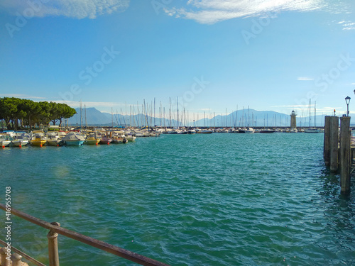 Lake landscape with sailboats, lighthouse, blue water, white clouds, mountains in haze. Lake Garda in Italy. Vacation concept