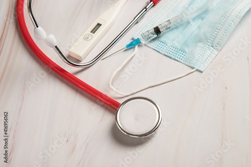A stethoscope, a syringe with a coronavirus vaccine, a medical mask, and an electronic thermometer on a light table. The concept of coronavirus vaccination. Copy space