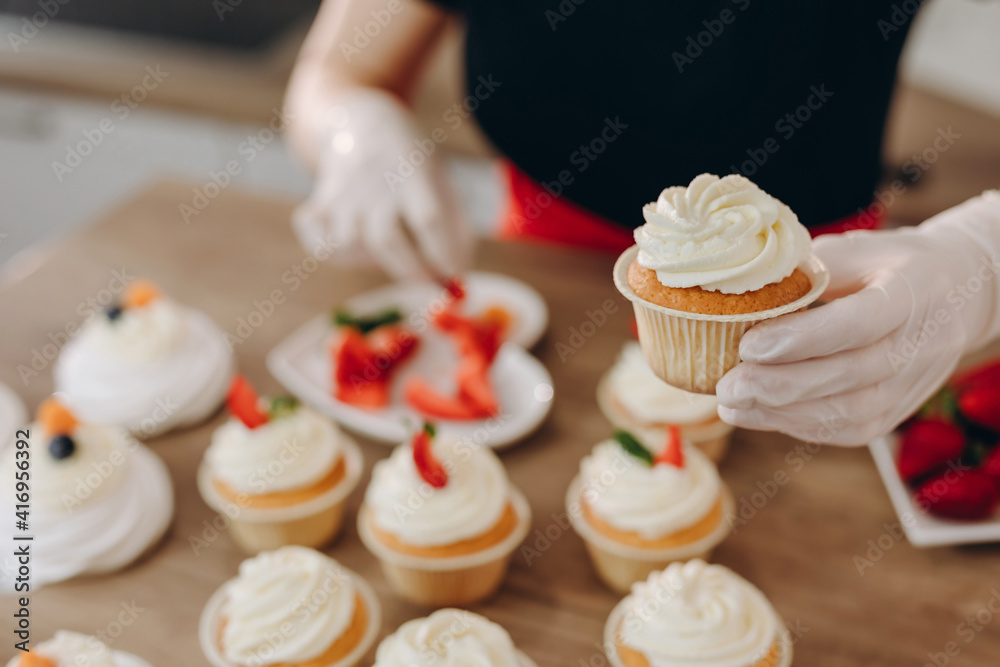 Close shot of many sweet cupcakes on the foreground while a baker decorating the last one.