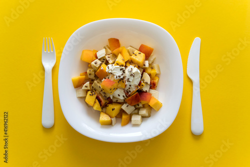 Bowl of healthy salad from fresh fruit on yellow. Healthy nutrition fruit salad with soft cheese and flax seeds in white plate on yellow background with white knife and fork.Healthy organic food.