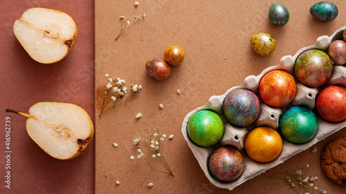 Happy Easter 2021. Colored painted eggs in a wicker basket on a brown background. Easter eggs and sweets. Cookies, pears, and chocolate.