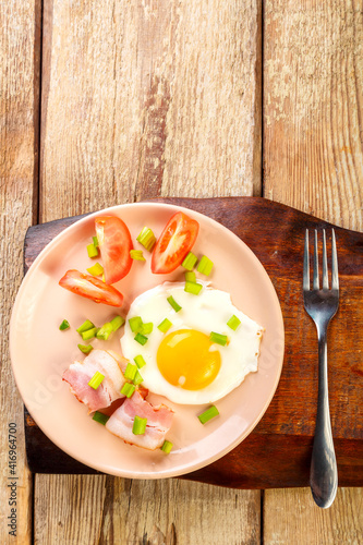 Scrambled eggs with bacon with tomato and onion and on a wooden table in a plate with a fork on a stand.