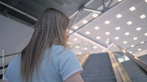 Slow motion: back view of young woman moving on escalator and looking around at mall, airoport or subway station - low angle photo