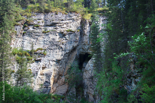 A huge rock among the trees in the Turgen Gorge, a stone gray sheer cliff with a cave in the middle, there are bushes on the rock here and there, summer, sunny