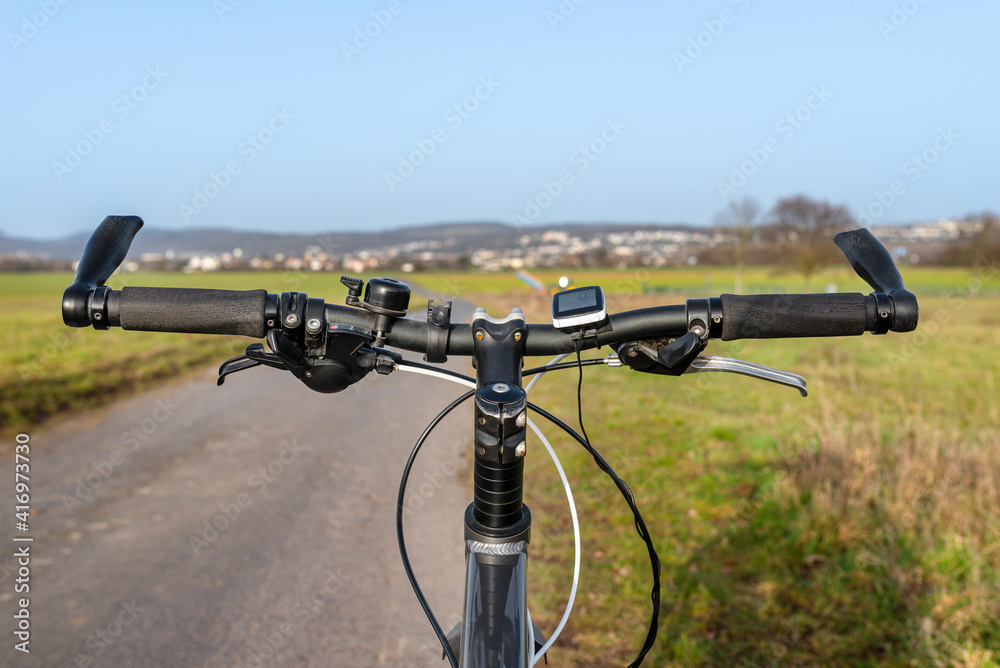 Mountain bike handlebar seen from the first person perspective. Visible bicycle frame and bicycle accessories on the handlebar, and the road in the background.