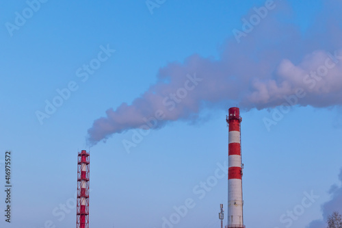 red and white chimney of a factory, from which gray smoke comes out against a blue sky on a sunny frosty winter day
