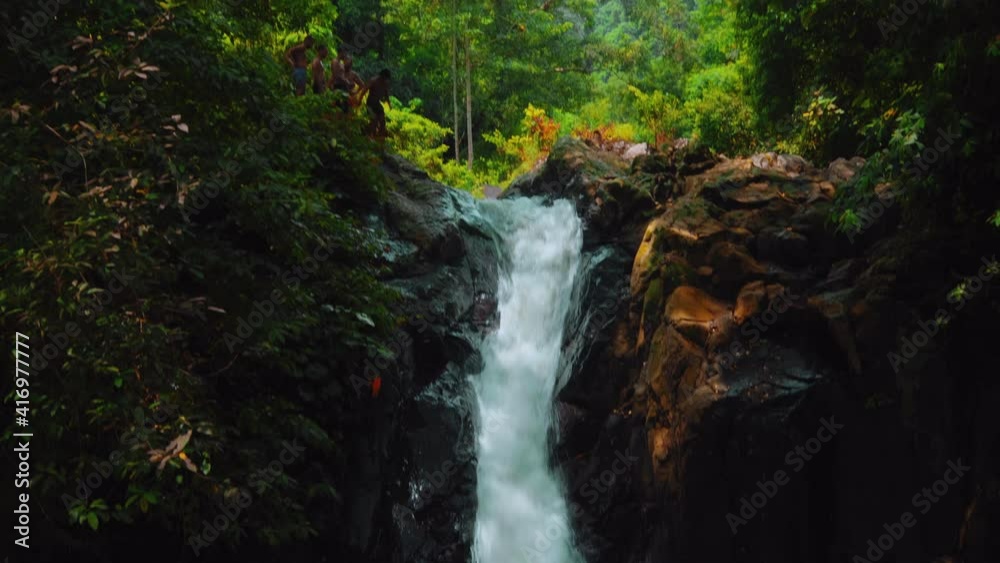 waterfall with cliffs among tropical jungle with green plants and trees and water falling down into the river with people standing on top faraway waterfall with cliffs among tropical jungle with green plants and trees and water falling down into the river with people standing on top faraway
