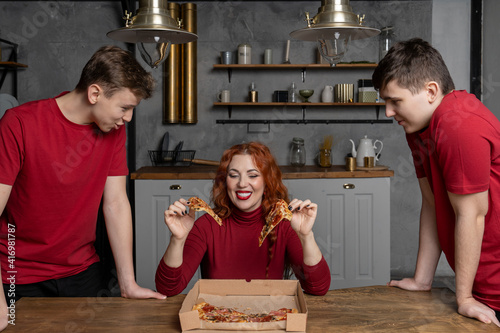 a woman sitting at the table with a smile holds two pieces of pizza in her hands and wants to give them to her teenage sons who are standing next to her