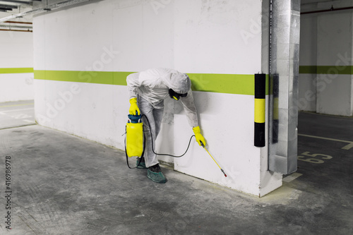 Manual worker in protective work wear spraying virus and bacteria disinfection in underground garage of residential building with sprayer.