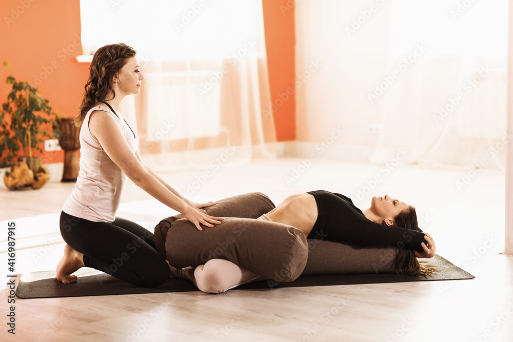Two women in sportswear practicing yoga doing Supta baddha konasana exercise with bolsters on a gymnastic mat in the studio Two women in sportswear practicing yoga doing Supta baddha konasana exercise with bolsters on a gymnastic mat in the studio