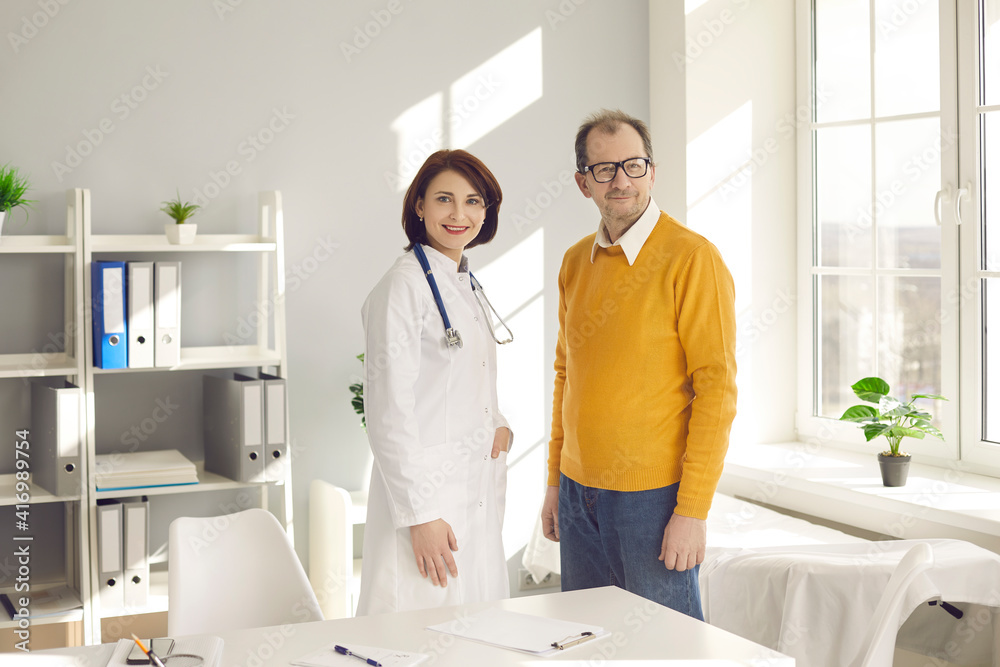 Fototapeta premium Portrait of happy female doctor and senior male patient in office of modern clinic. Smiling mature man and general practitioner standing together and looking at camera. Health screening visit concept