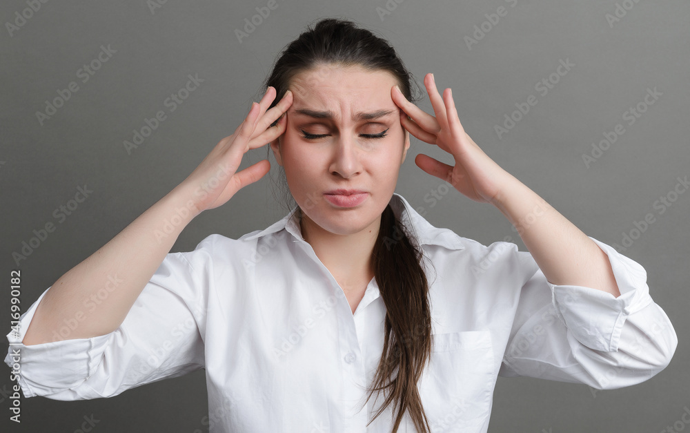 1 white young woman in a white shirt holding her head with her hands on a gray , headache, 1 white young woman in a white shirt holding her head with her hands on a gray , headache,