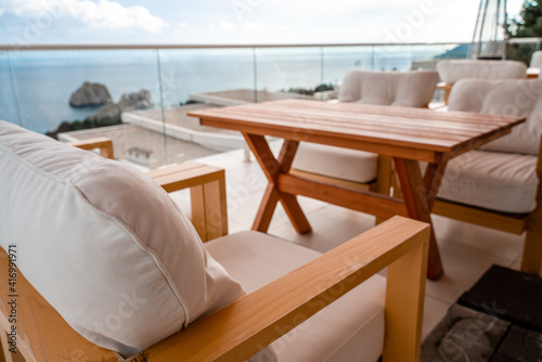 Restaurant with sea view. You can see rocks in the sea. Wooden tables and chairs with white cushions. Overcast  cloudy weather.