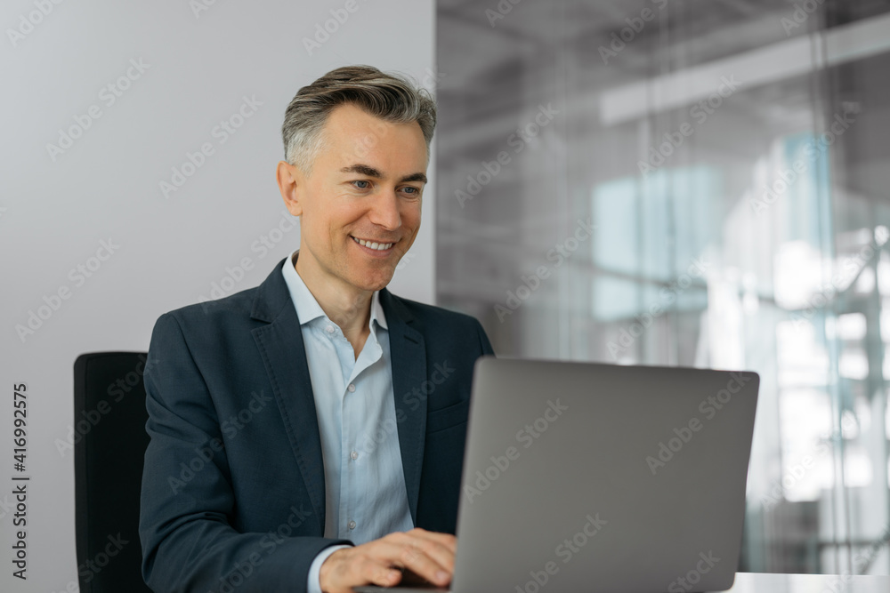 Handsome mature businessman using laptop computer working online sitting in office. Portrait of successful smiling programmer at workplace Handsome mature businessman using laptop computer working online sitting in office. Portrait of successful smiling programmer at workplace