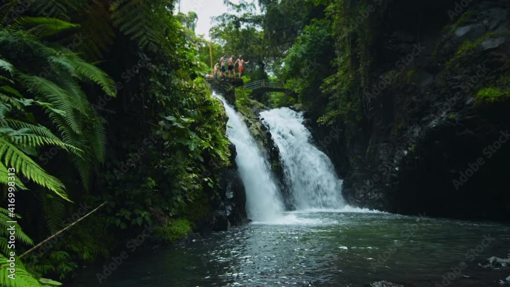 waterfall with cliffs among tropical jungle with green plants and trees and water falling down into the river with people standing on top faraway waterfall with cliffs among tropical jungle with green plants and trees and water falling down into the river with people standing on top faraway