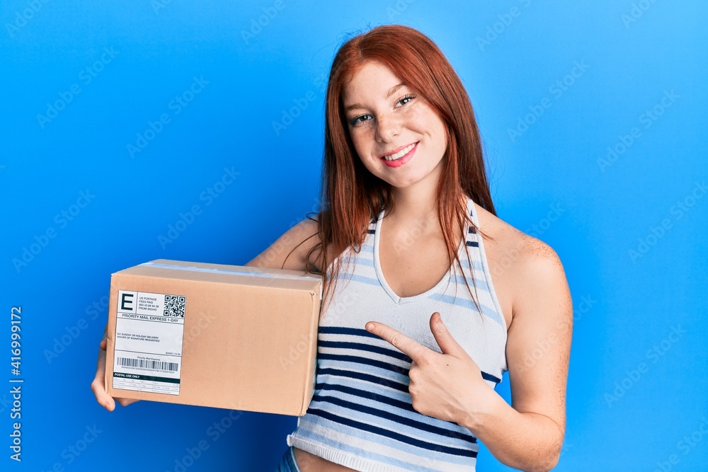 Young red head girl holding delivery package smiling happy pointing with hand and finger Young red head girl holding delivery package smiling happy pointing with hand and finger