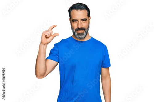 Middle aged man with beard wearing casual blue t shirt smiling and confident gesturing with hand doing small size sign with fingers looking and the camera. measure concept.