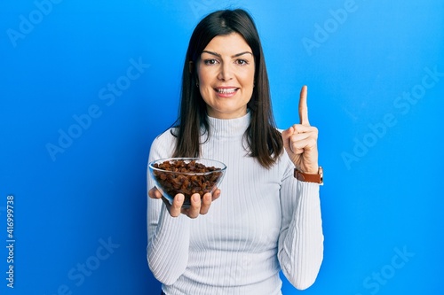 Young hispanic woman holding raisins bowl smiling with an idea or question pointing finger with happy face  number one