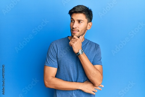 Young handsome man wearing casual tshirt over blue background thinking concentrated about doubt with finger on chin and looking up wondering