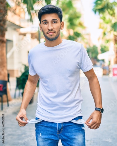 Young latin man with worried expression showing empty pockets walking at the city.