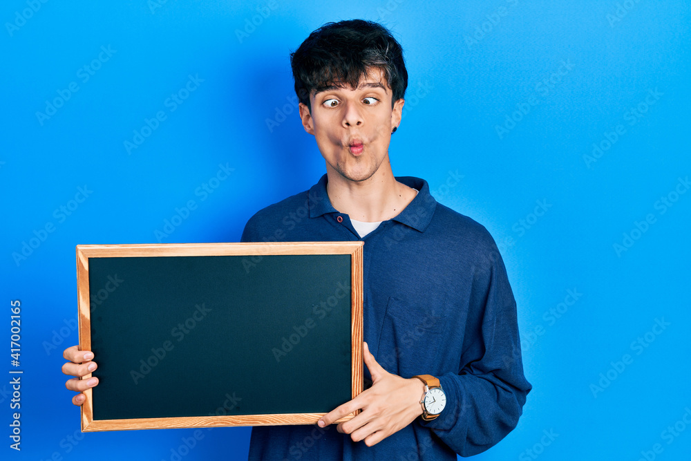 Handsome hipster young man holding blackboard making fish face with mouth and squinting eyes, crazy and comical. Handsome hipster young man holding blackboard making fish face with mouth and squinting eyes, crazy and comical.