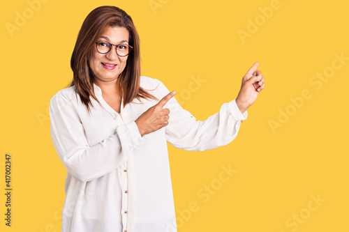 Middle age latin woman wearing casual clothes and glasses smiling and looking at the camera pointing with two hands and fingers to the side.