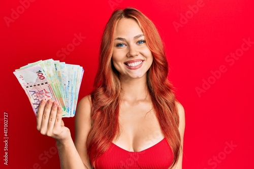 Young redhead woman holding hong kong dollars banknotes looking positive and happy standing and smiling with a confident smile showing teeth