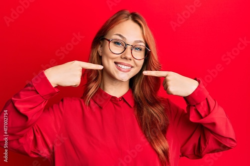 Young beautiful redhead woman wearing casual clothes and glasses over red background smiling cheerful showing and pointing with fingers teeth and mouth. dental health concept.