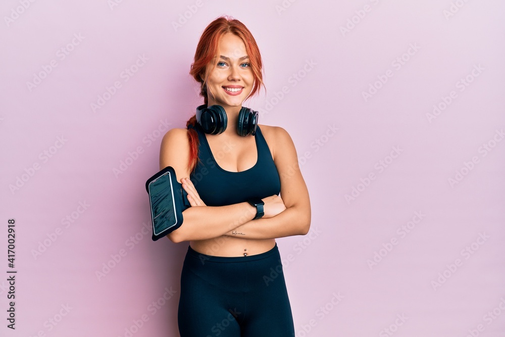 Young beautiful redhead woman wearing gym clothes and using headphones happy face smiling with crossed arms looking at the camera. positive person.