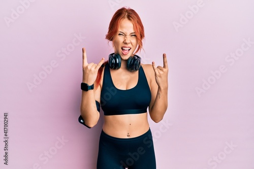 Young beautiful redhead woman wearing gym clothes and using headphones shouting with crazy expression doing rock symbol with hands up. music star. heavy concept.
