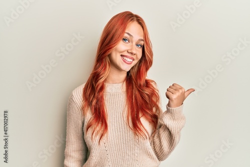 Young beautiful redhead woman wearing casual winter sweater smiling with happy face looking and pointing to the side with thumb up.