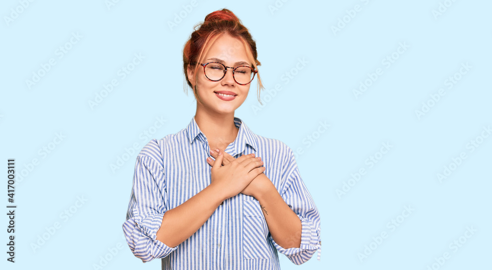 Young redhead woman wearing casual clothes and glasses smiling with hands on chest with closed eyes and grateful gesture on face. health concept.