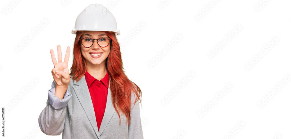 Young redhead woman wearing architect hardhat showing and pointing up with fingers number three while smiling confident and happy.