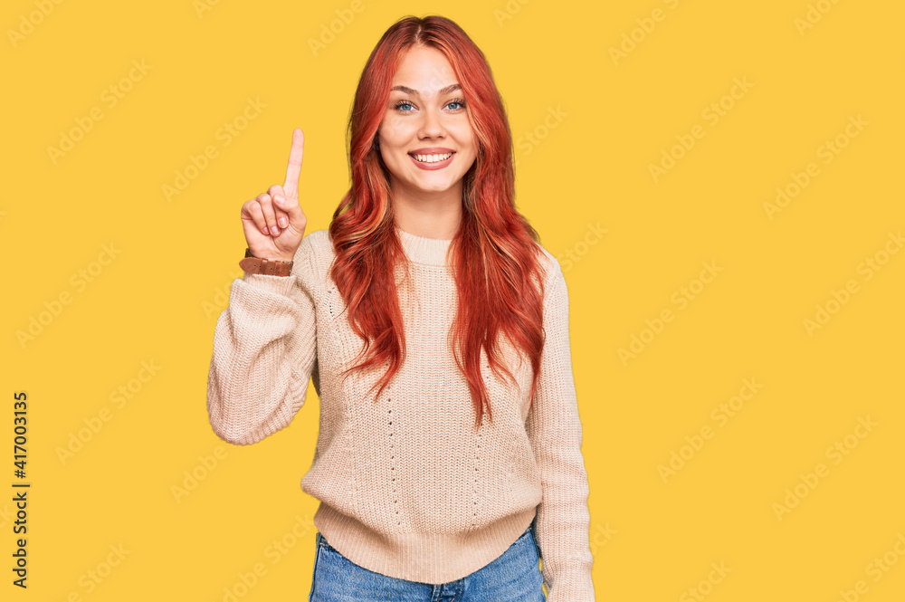 Young redhead woman wearing casual winter sweater showing and pointing up with finger number one while smiling confident and happy. Young redhead woman wearing casual winter sweater showing and pointing up with finger number one while smiling confident and happy.