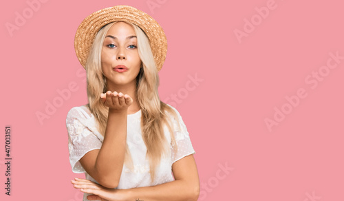 Young blonde girl wearing summer hat looking at the camera blowing a kiss with hand on air being lovely and sexy. love expression.