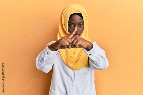 Beautiful african young woman wearing doctor uniform and hijab rejection expression crossing fingers doing negative sign