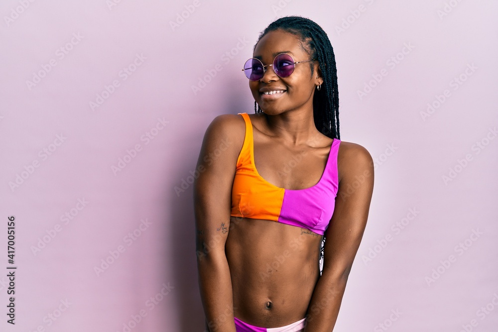 Naklejka premium Young african american woman wearing bikini and sunglasses smiling looking to the side and staring away thinking.