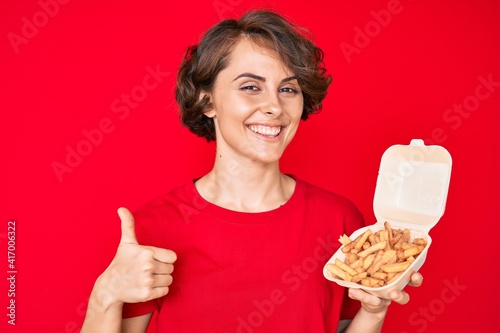 Young hispanic woman holding potato chip smiling happy and positive  thumb up doing excellent and approval sign