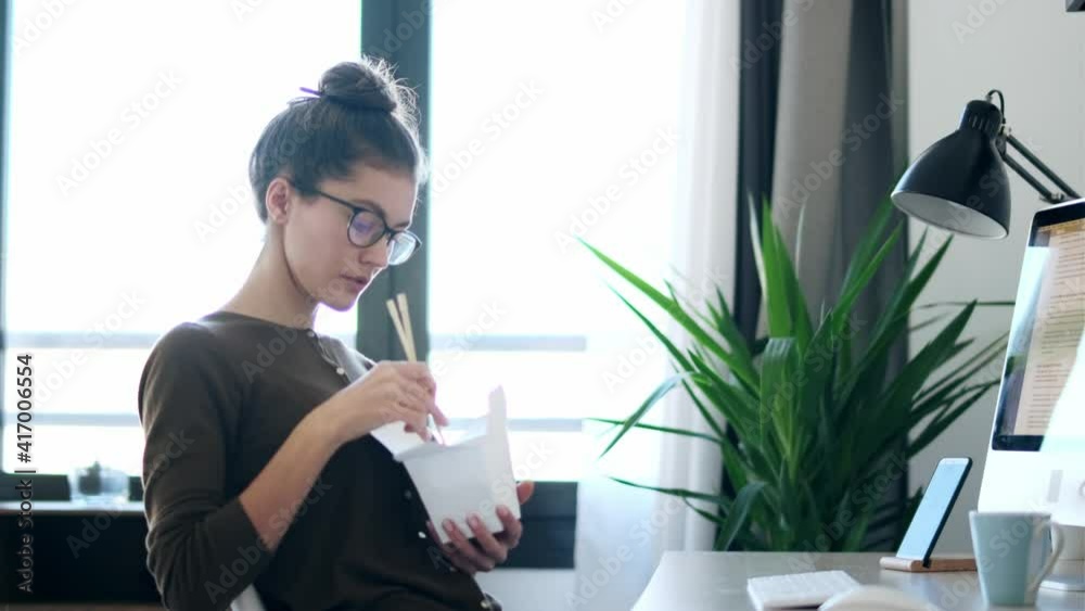 Video of happy young woman eating noodles with chopsticks while taking a break of work sitting on chair in the office at home.