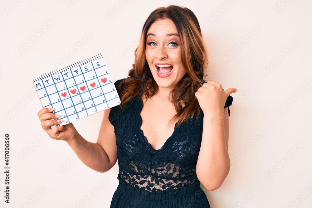 Young beautiful caucasian woman holding heart calendar pointing thumb up to the side smiling happy with open mouth Young beautiful caucasian woman holding heart calendar pointing thumb up to the side smiling happy with open mouth