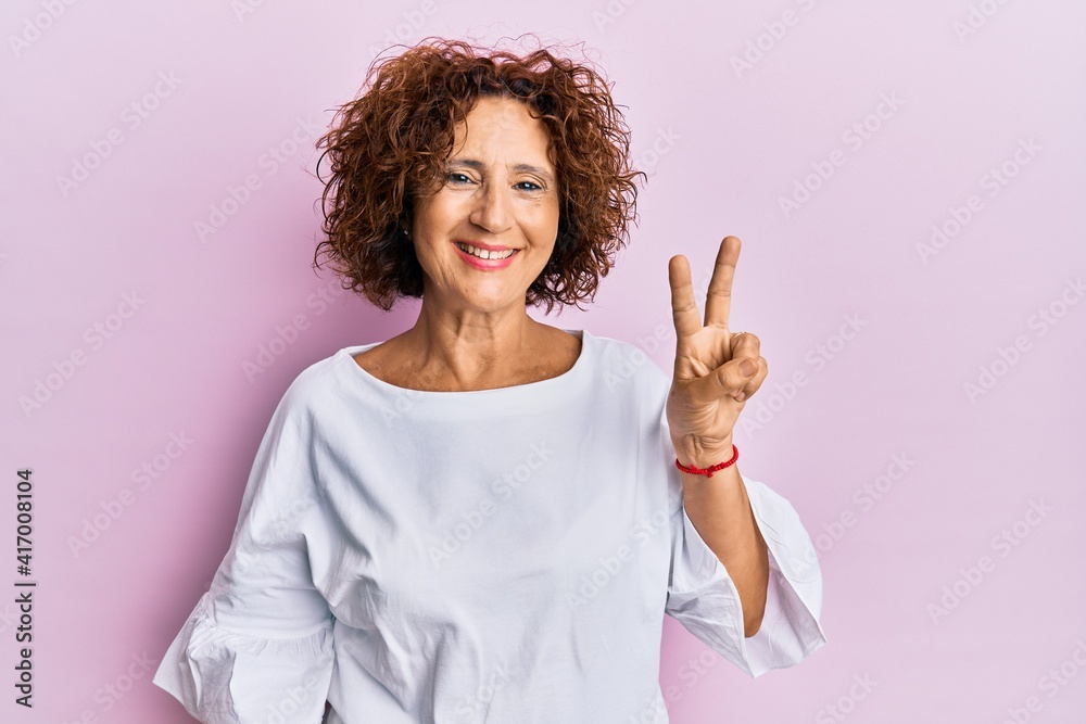 Beautiful middle age mature woman wearing elegant clothes over pink background smiling with happy face winking at the camera doing victory sign. number two. Beautiful middle age mature woman wearing elegant clothes over pink background smiling with happy face winking at the camera doing victory sign. number two.