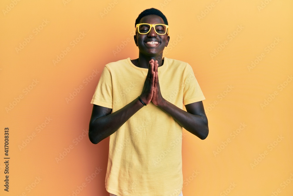 Young african american man wearing casual clothes and glasses praying with hands together asking for forgiveness smiling confident. Young african american man wearing casual clothes and glasses praying with hands together asking for forgiveness smiling confident.