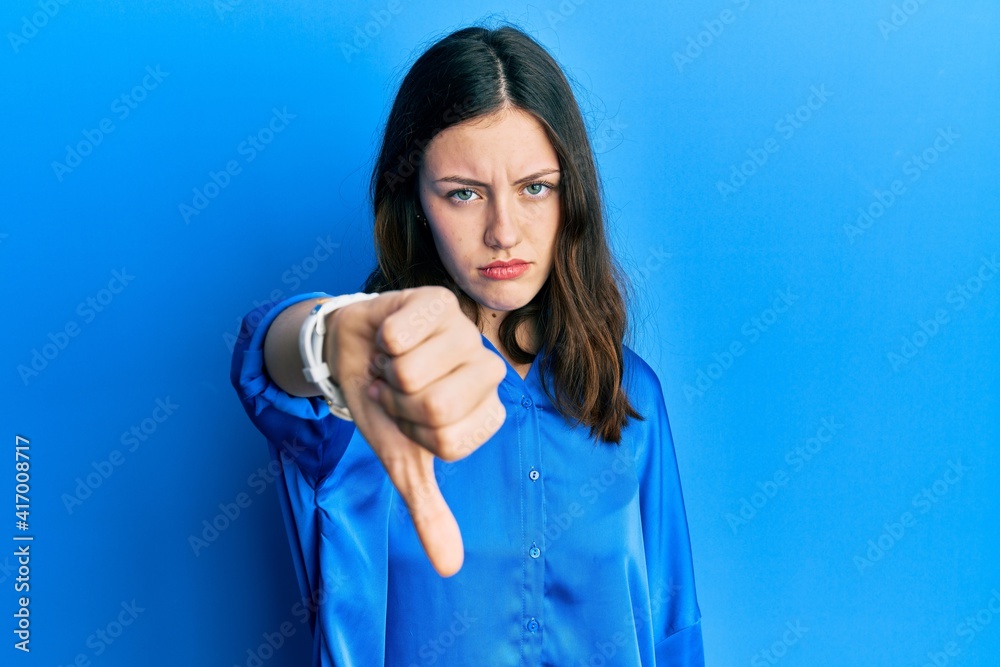 Young brunette woman wearing casual blue shirt looking unhappy and angry showing rejection and negative with thumbs down gesture. bad expression. Young brunette woman wearing casual blue shirt looking unhappy and angry showing rejection and negative with thumbs down gesture. bad expression.