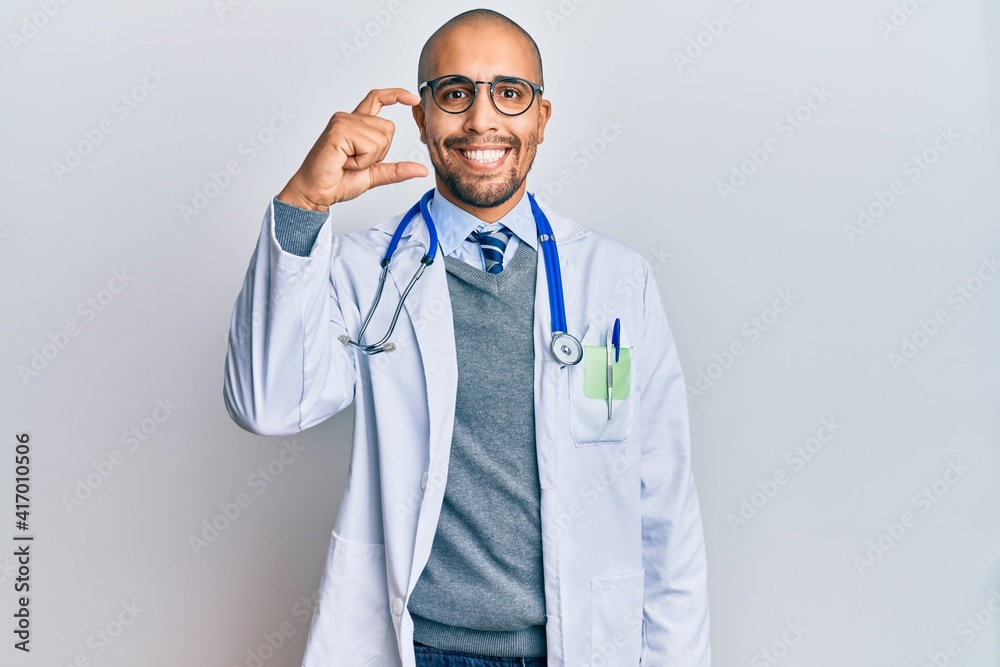 Hispanic adult man wearing doctor uniform and stethoscope smiling and confident gesturing with hand doing small size sign with fingers looking and the camera. measure concept. Hispanic adult man wearing doctor uniform and stethoscope smiling and confident gesturing with hand doing small size sign with fingers looking and the camera. measure concept.
