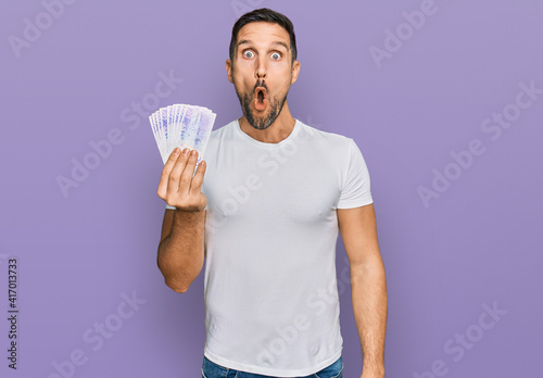 Handsome man with beard holding 20 swedish krona banknotes scared and amazed with open mouth for surprise, disbelief face