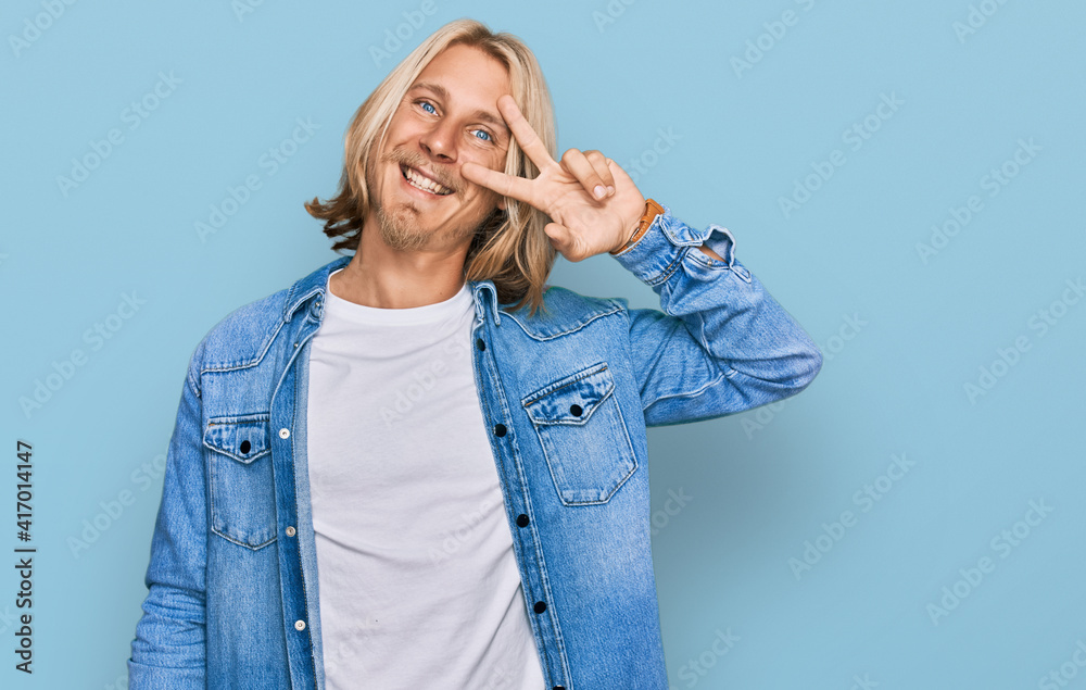 Caucasian man with blond long hair wearing casual denim jacket doing peace symbol with fingers over face, smiling cheerful showing victory Caucasian man with blond long hair wearing casual denim jacket doing peace symbol with fingers over face, smiling cheerful showing victory