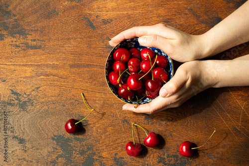hands holding bowl with cherries on dark wooden table, top view 