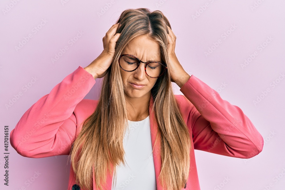 Young blonde woman wearing business style and glasses suffering from headache desperate and stressed because pain and migraine. hands on head. Young blonde woman wearing business style and glasses suffering from headache desperate and stressed because pain and migraine. hands on head.