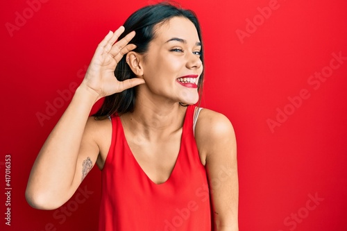 Young hispanic girl wearing casual style with sleeveless shirt smiling with hand over ear listening an hearing to rumor or gossip. deafness concept.