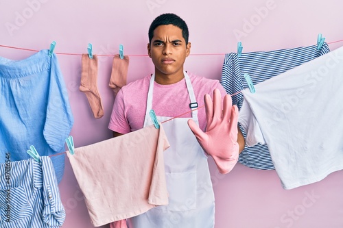 Young handsome hispanic man wearing cleaner apron holding clothes on clothesline doing stop sing with palm of the hand. warning expression with negative and serious gesture on the face.